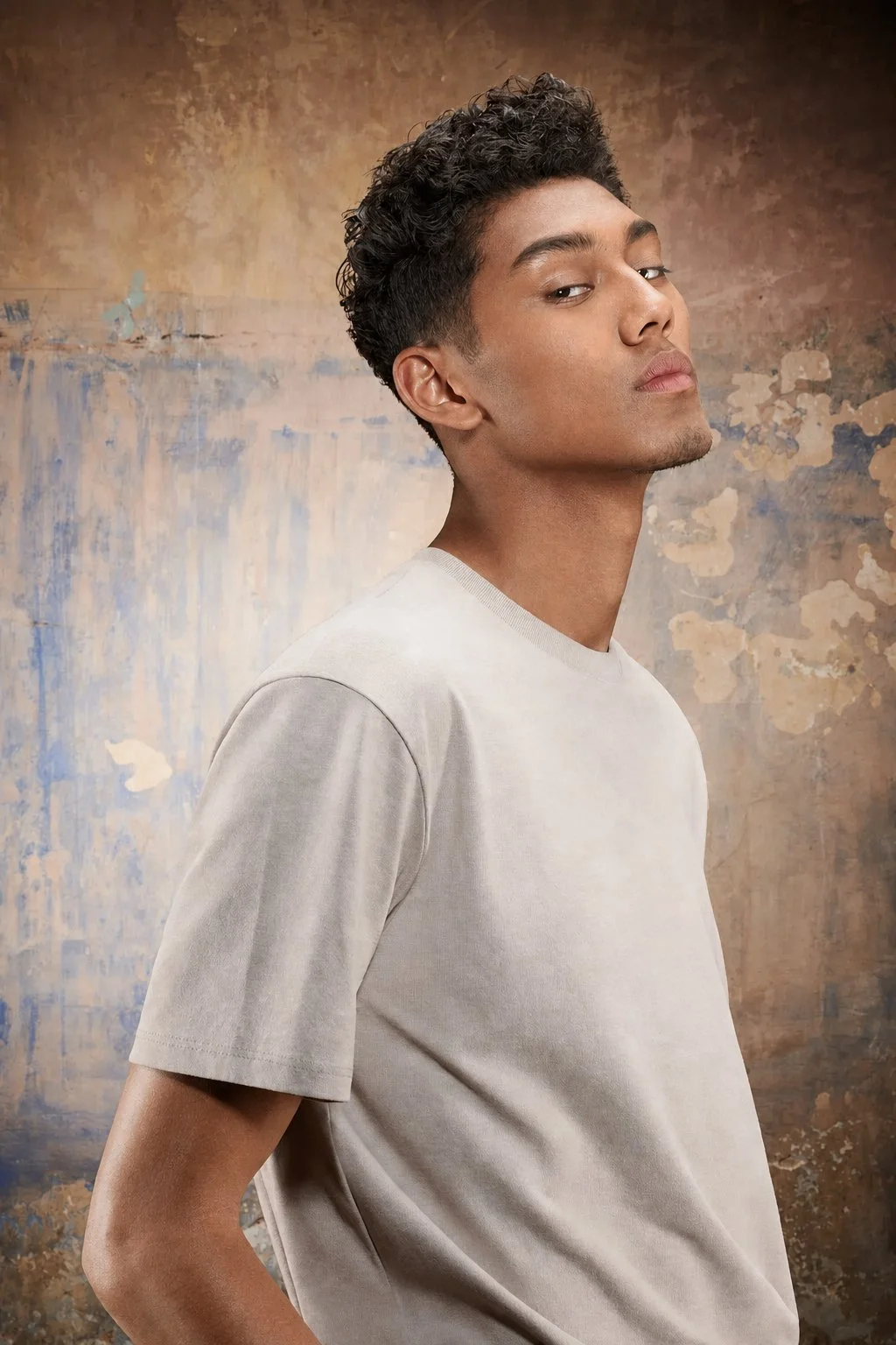 Young man with dark curly hair and medium skin tone, wearing a light-colored t-shirt, standing in front of a textured, abstract brown and beige background, looking over his shoulder with a confident expression.