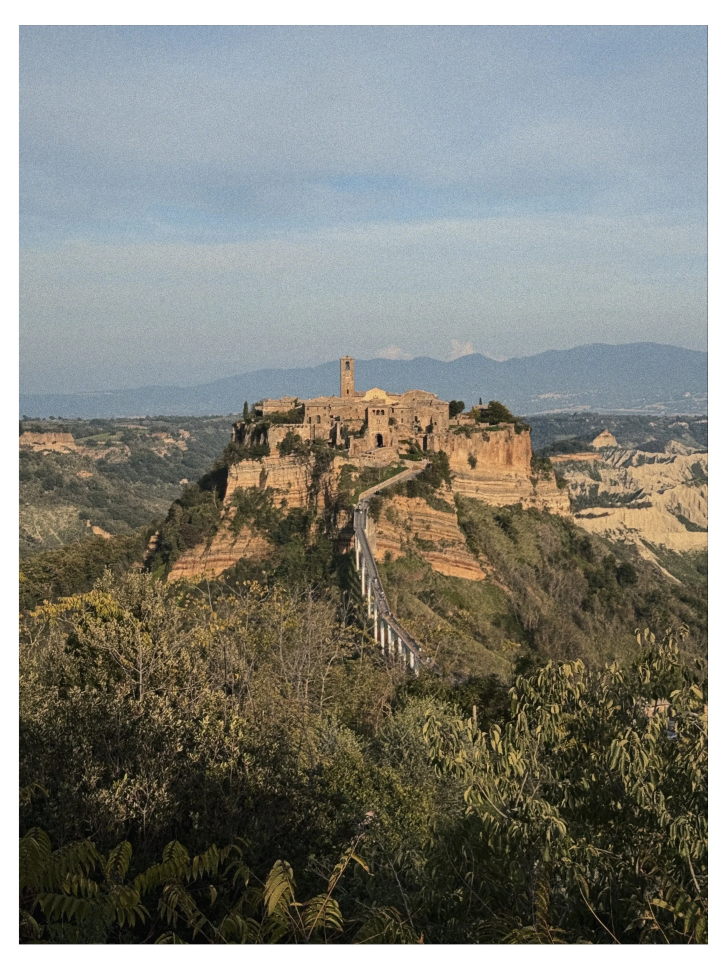 A hilltop town with ancient stone buildings and a tall, narrow bell tower, perched on a rugged cliff overlooking a scenic landscape of green hills and distant mountains, under a partly cloudy sky.