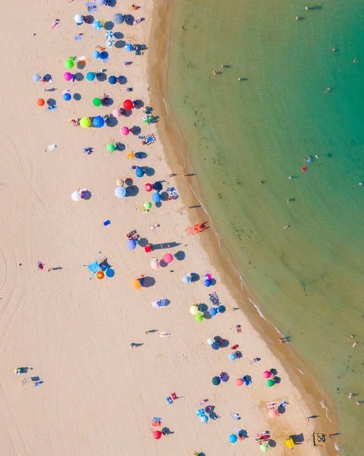 Aerial view of a crowded beach with colorful umbrellas and people relaxing on the sand and swimming in the water.