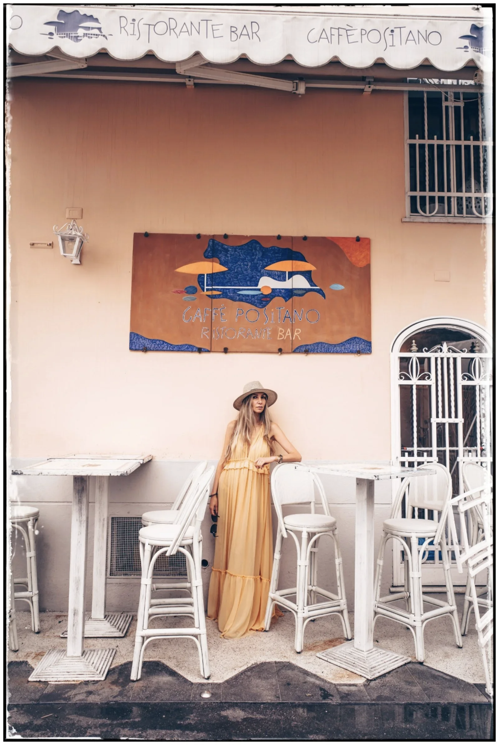 A young woman in a long yellow dress and wide-brimmed straw hat standing outside a cafe with a peach-colored wall. There are white tables and chairs, a sign overhead reading 'Ristorante Bar Caffé Italiano,' and a decorative sign behind her with a blu