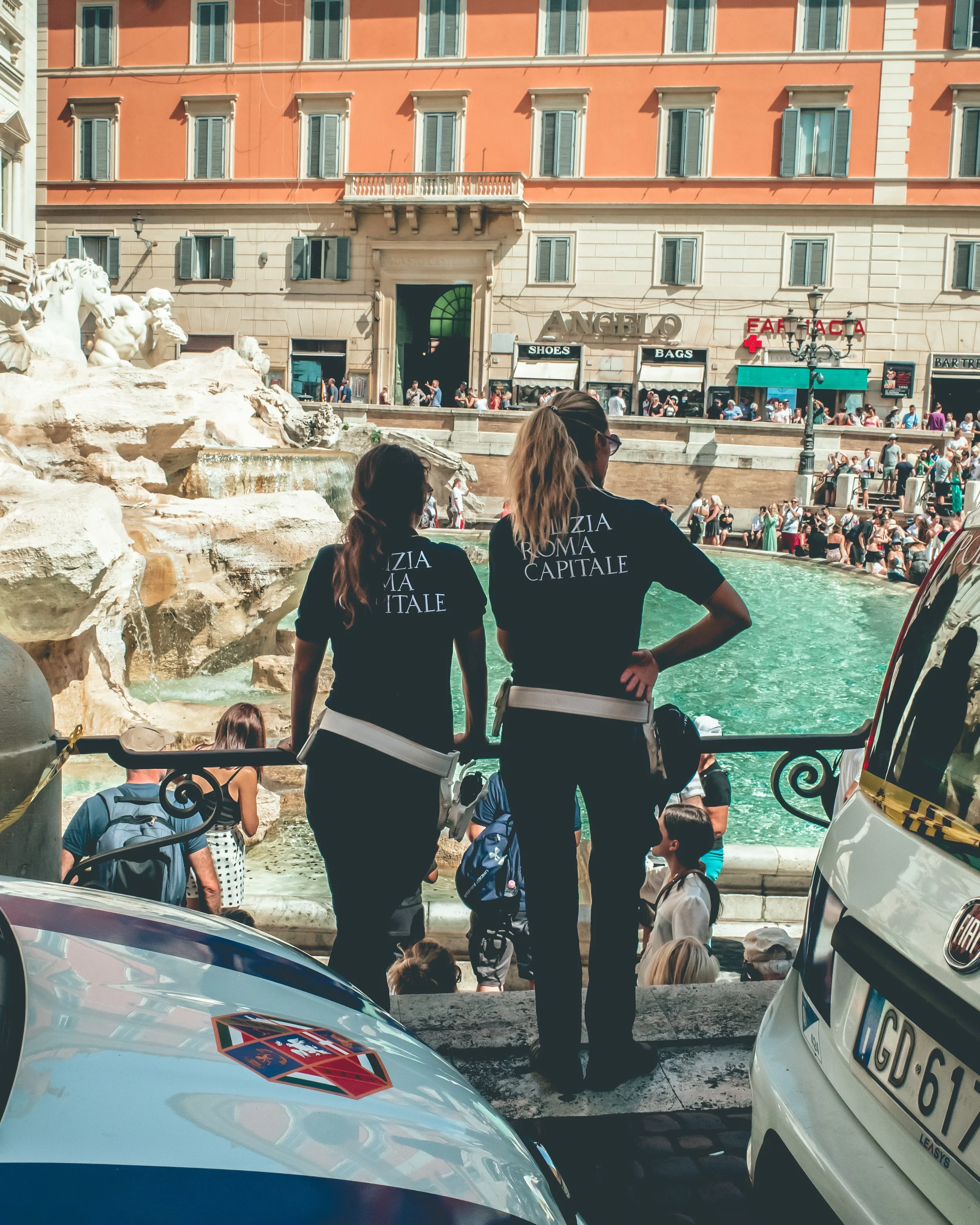 People gather near a fountain in a busy city square with historic buildings, shops, and a pharmacy. Two women in black shirts with white lettering stand in the foreground, overlooking the fountain, while many other people walk or stand around.