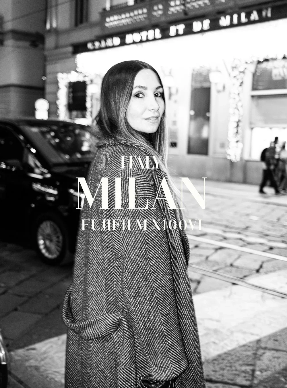 A young woman with long hair standing on a city street at night, wearing a patterned coat, with cars and a lit building in the background.