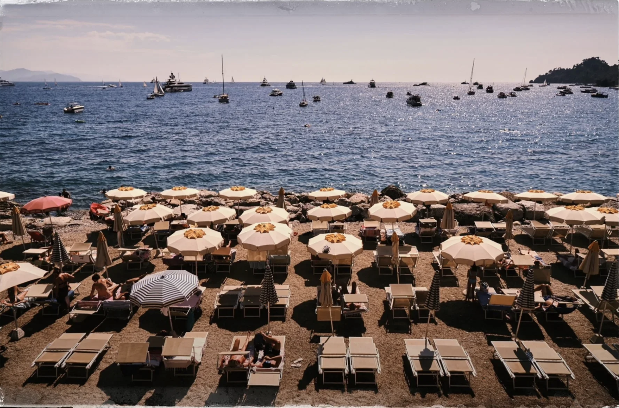 Beach scene with rows of lounge chairs and umbrellas on a sandy shore, with people sunbathing and relaxing, and boats anchored on the calm water in the background under a clear sky.
