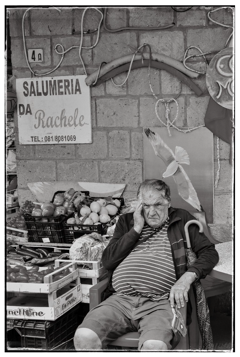 An elderly man with gray hair, wearing a striped shirt and shorts, sits on a plastic chair in a market. He rests his head on his hand with a frustrated or tired expression. Behind him are crates of vegetables, and a sign that reads 'Salumeria da Rach