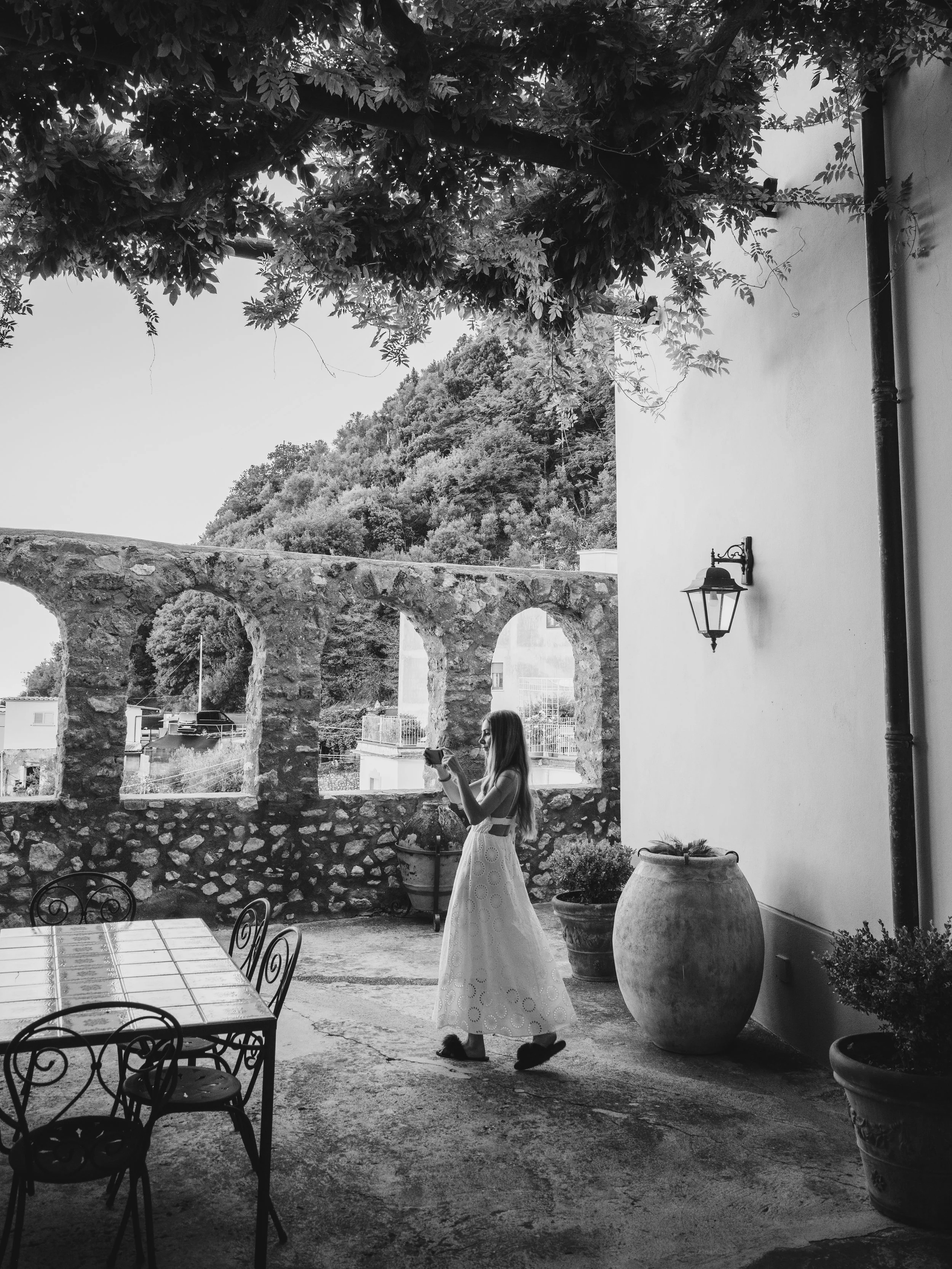 A woman in a white dress with eyelet details stands outdoors on a patio, looking at her phone, while a dog sits beside her. The patio is decorated with large potted plants, metal chairs, and tables. In the background, there is an old stone archway wi