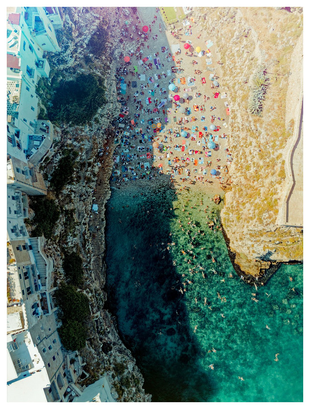 Bird's eye view of a crowded beach with colorful umbrellas and people swimming in the turquoise water, surrounded by rocky cliffs and white buildings.