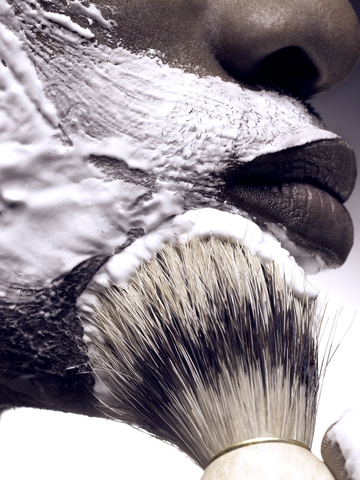 Close-up of a shaving brush with a person's face and beard covered in white foam, preparing for a shave.