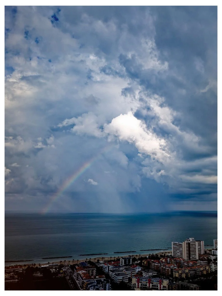 View of a cloudy sky with a faint rainbow over an ocean and cityscape with high-rise buildings.