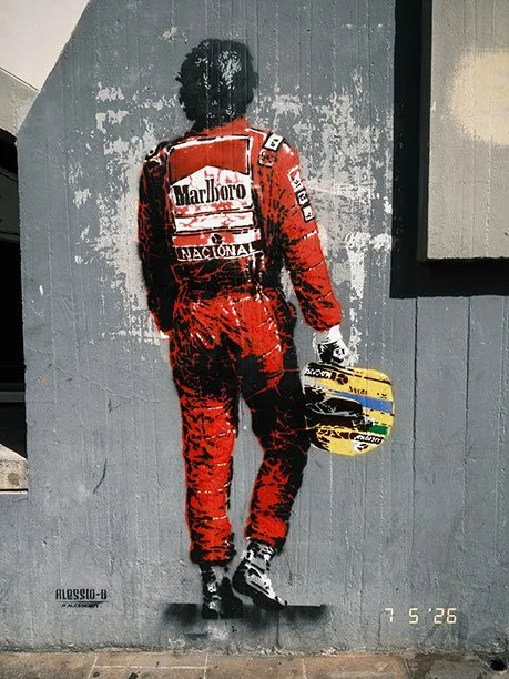 Street art mural of a man in a red racing suit holding a helmet, facing away, on a gray wall.