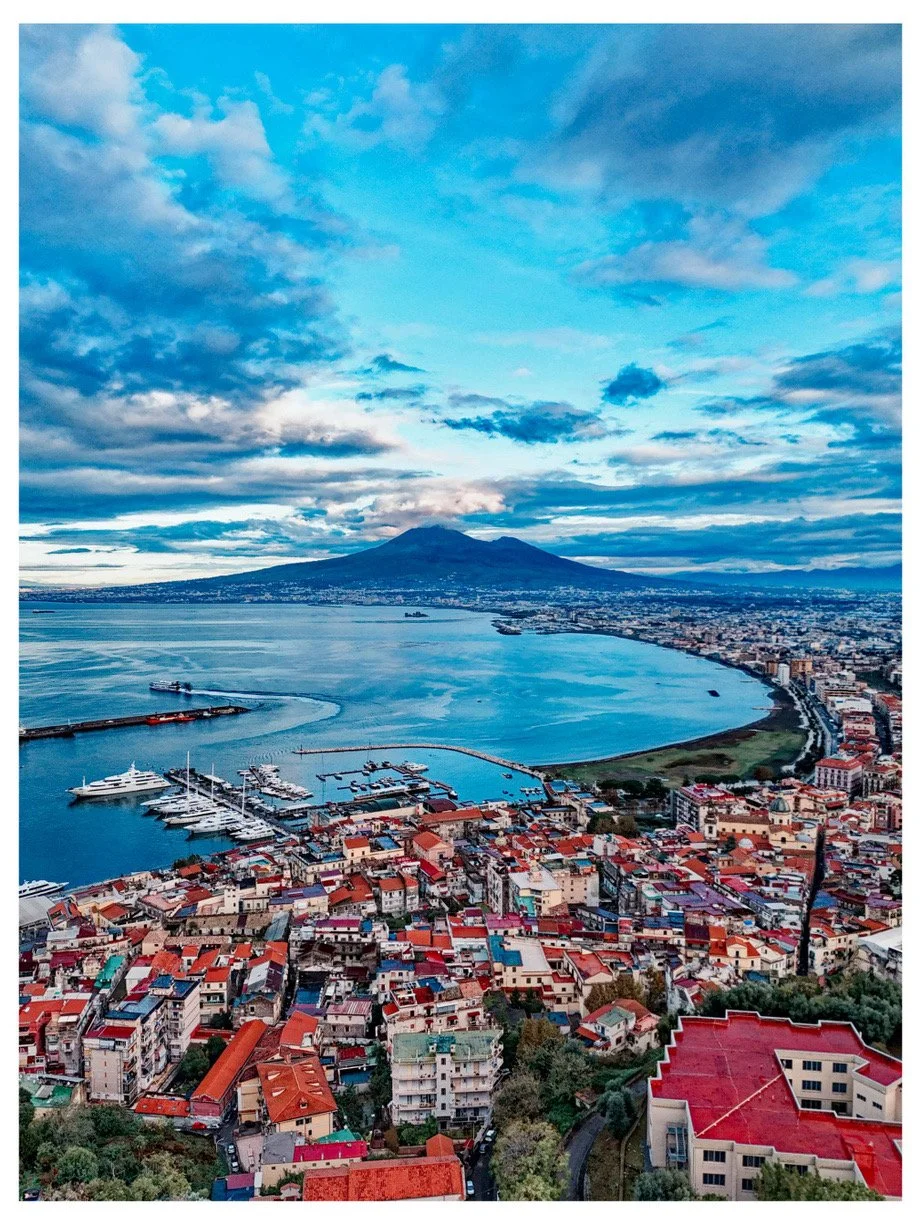 Aerial view of a coastal city with a harbor, boats, and a mountain in the background, under a cloudy blue sky.