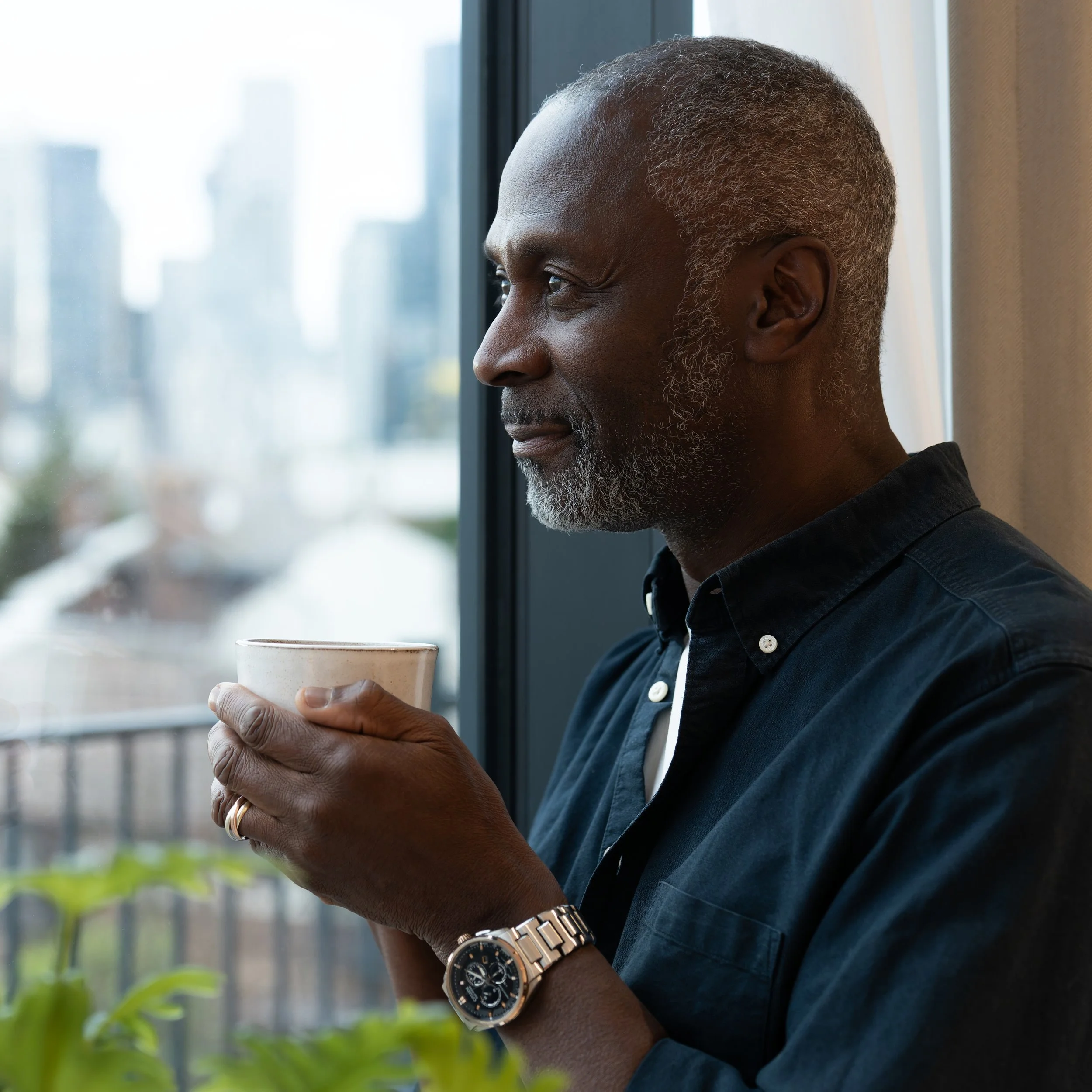 Close-up of an older African American man with gray hair, holding a white ceramic mug, sitting near a window with city buildings in the background.