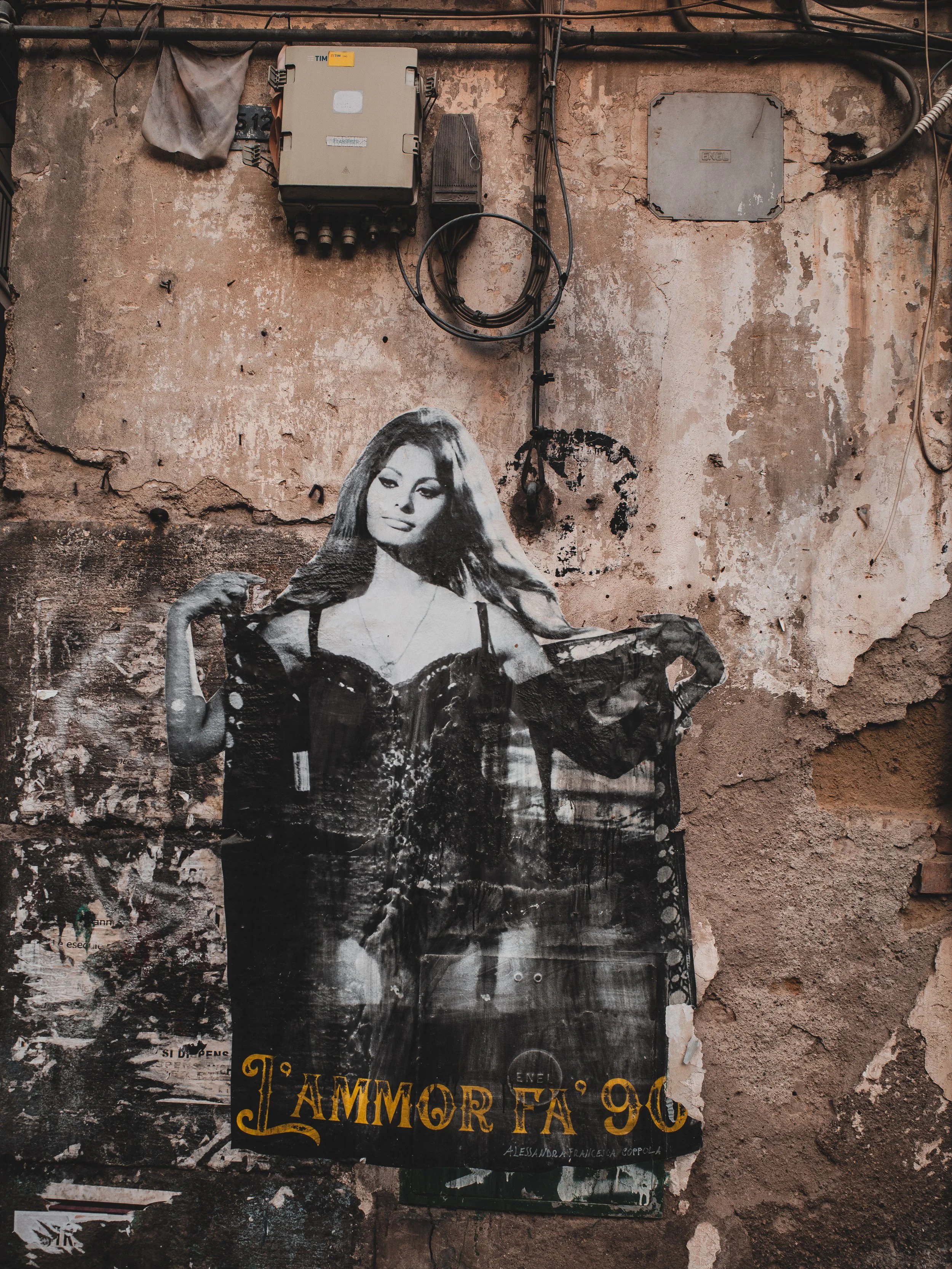 A black and white poster of a woman with long hair, posing with one arm flexed and the other resting on her hips, hanging on a weathered wall with peeling paint and electrical equipment above.