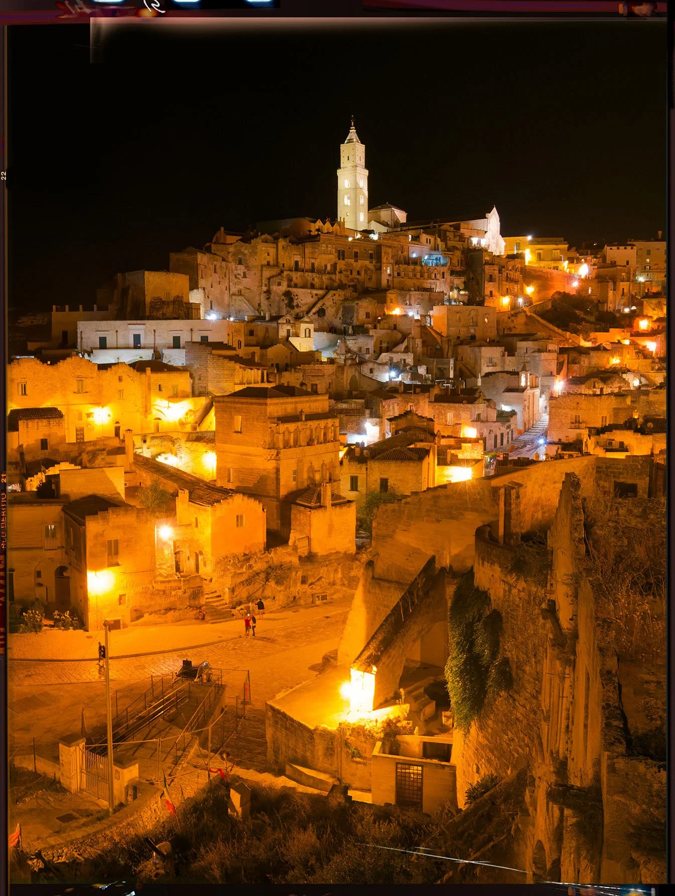 Night view of a historic hillside town illuminated by warm yellow lights, with a prominent tall church tower at the top of the hill and smaller buildings and steps winding down the slope.