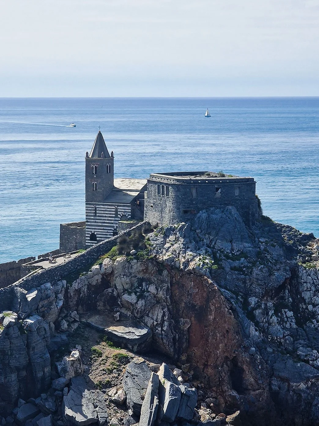A historic stone church with a tall steeple on a rocky coastline, overlooking the ocean with a sailboat and a motorboat in the distance.