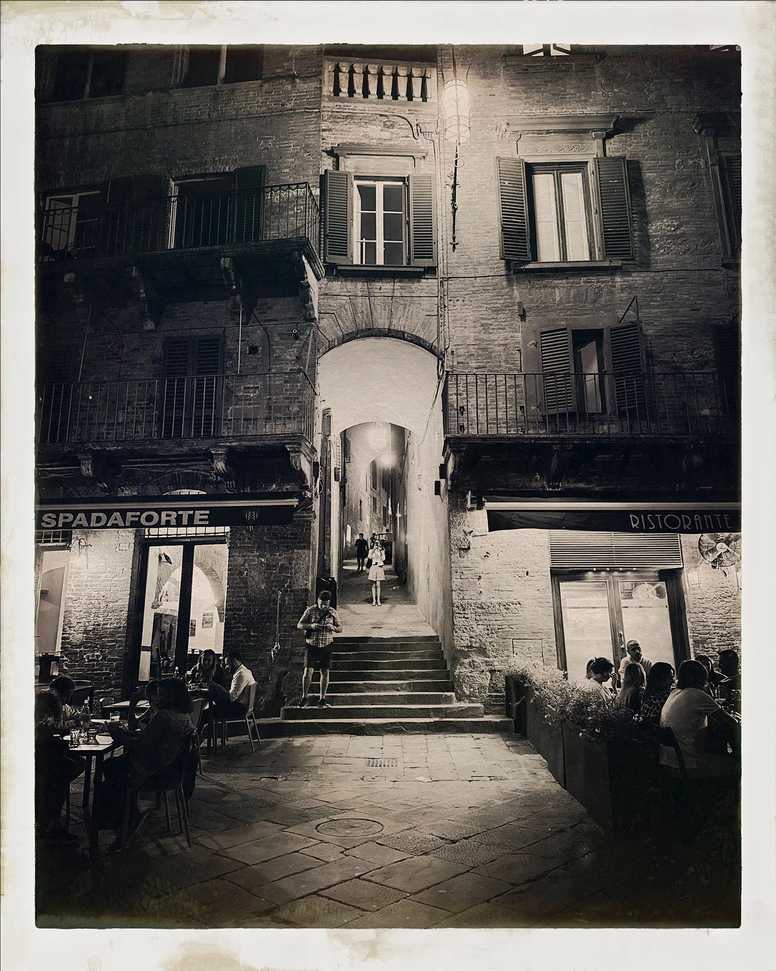 Night scene of a historic brick building with open shutter windows and balconies, a lit streetlight, and a narrow passageway arched beneath the building. People are dining and walking outside a restaurant with outdoor seating.