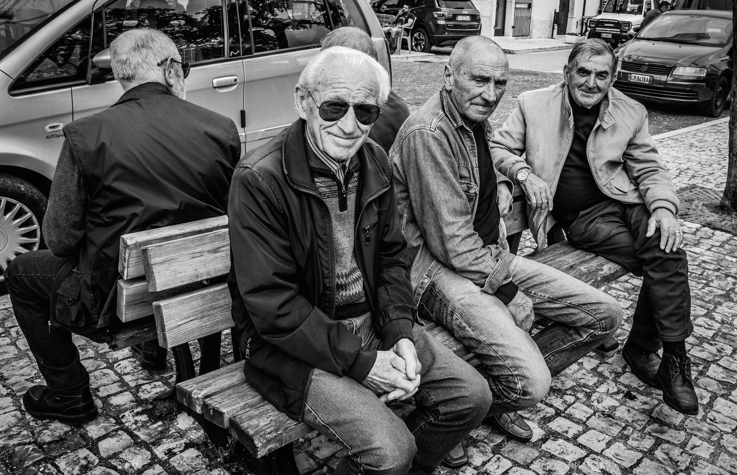 Four elderly men sitting on a wooden bench outdoors, some wearing jackets, in front of parked cars on a cobblestone street.