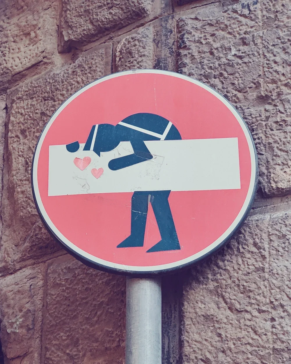 A street sign with a sticker of a person holding a white rectangle and blowing hearts, placed over the original sign image.