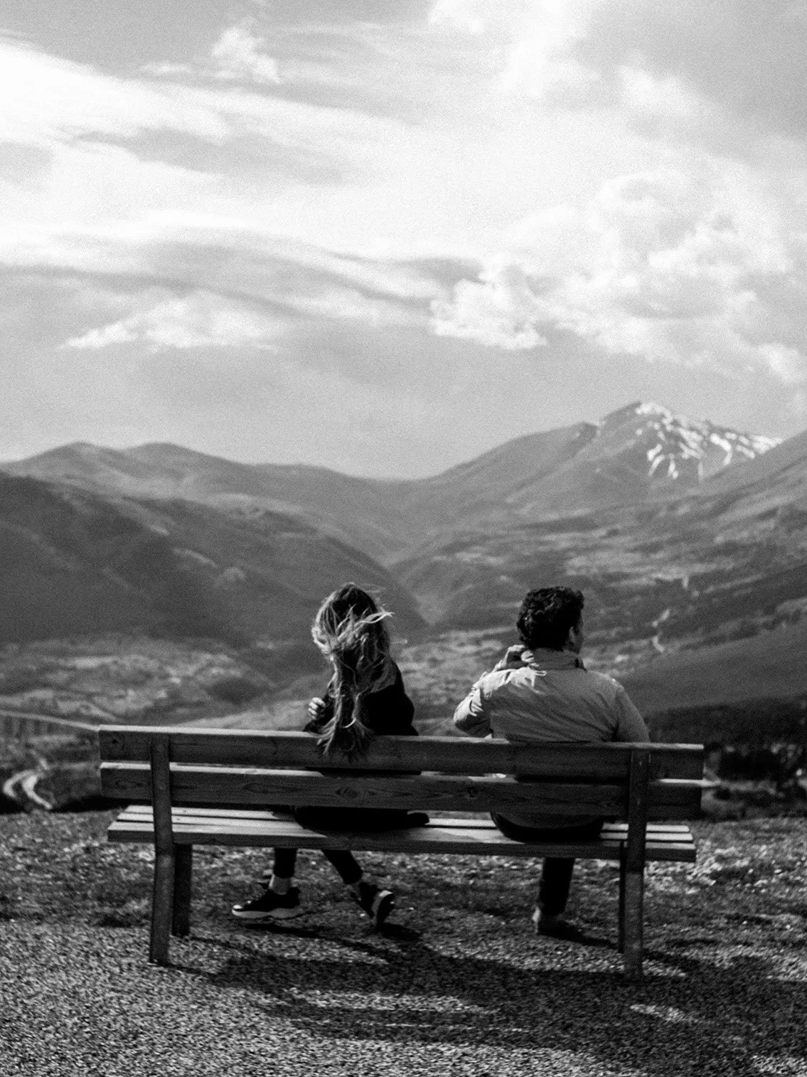 Two people, a man and a woman, sitting on a wooden bench outdoors, looking at a mountainous landscape with snow-capped peaks in the distance. The photo is in black and white.