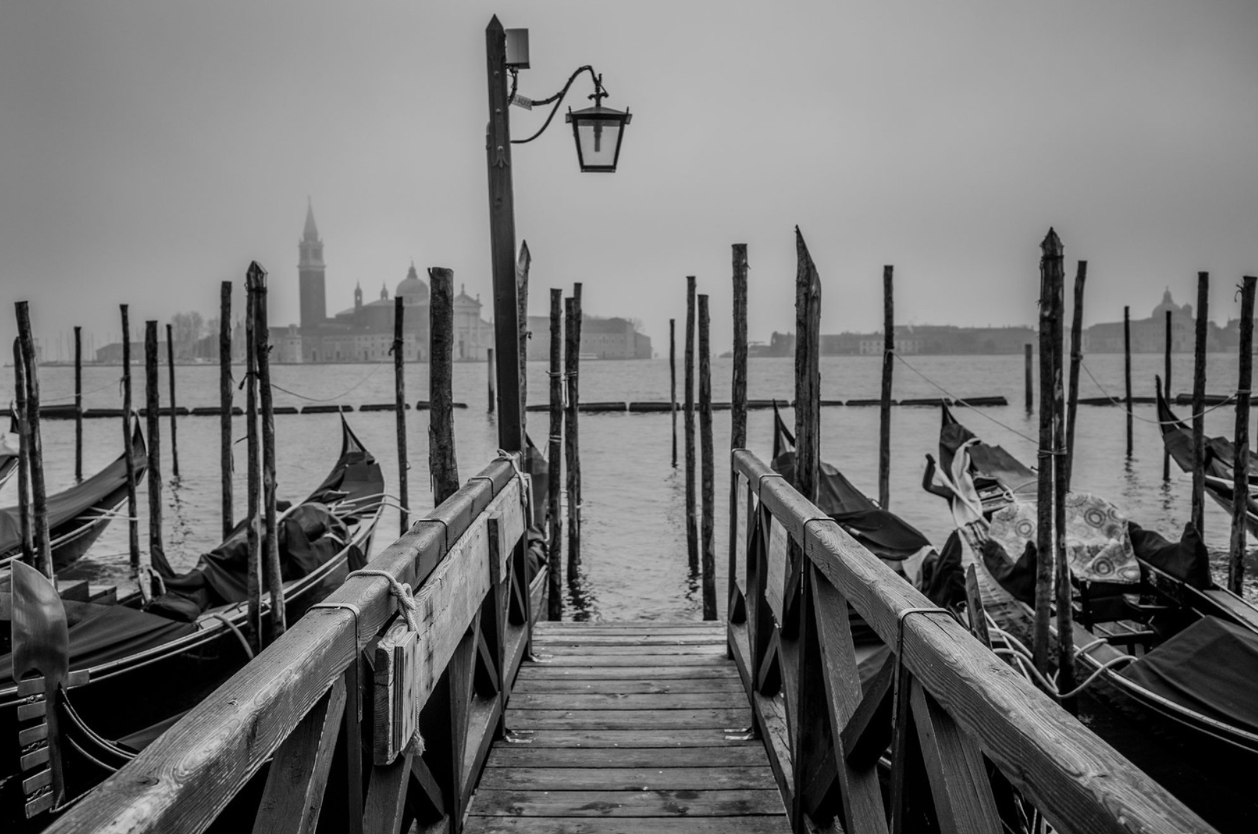 A black-and-white photo of Venice's waterfront with a wooden dock leading to gondolas tied to weathered wooden posts. A vintage street lamp hangs from a pole near the dock, with the city skyline and a tall bell tower in the foggy distance.
