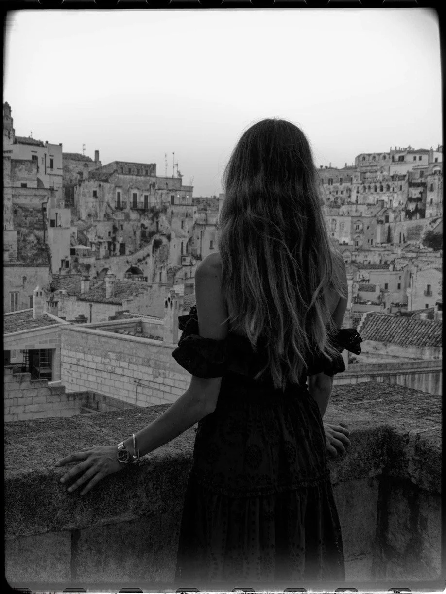 A woman with long hair standing on a balcony, overlooking a densely built cityscape with old stone buildings, in black and white.