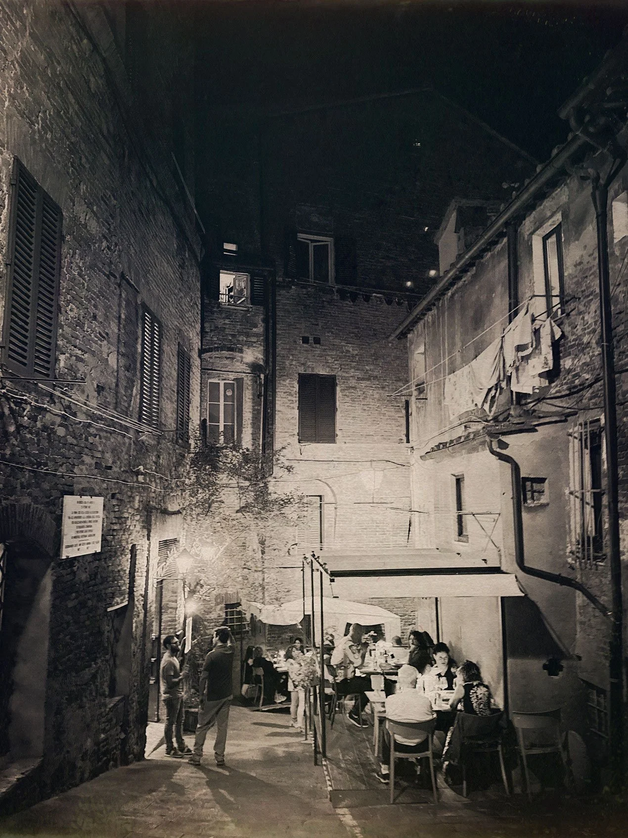 Nighttime outdoor scene of a narrow alley with brick buildings on either side, some windows with shutters, laundry hanging on a line, and people sitting at tables under an umbrella, illuminated by string lights.