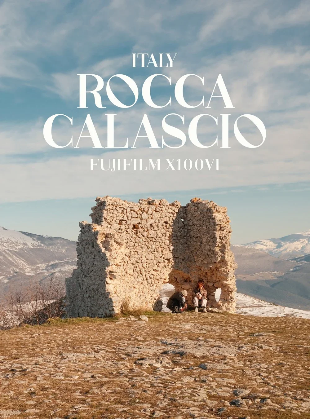 A scenic landscape with ruins of an ancient stone structure, snow-capped mountains in the background, and three people sitting near the ruins, under a partly cloudy sky.