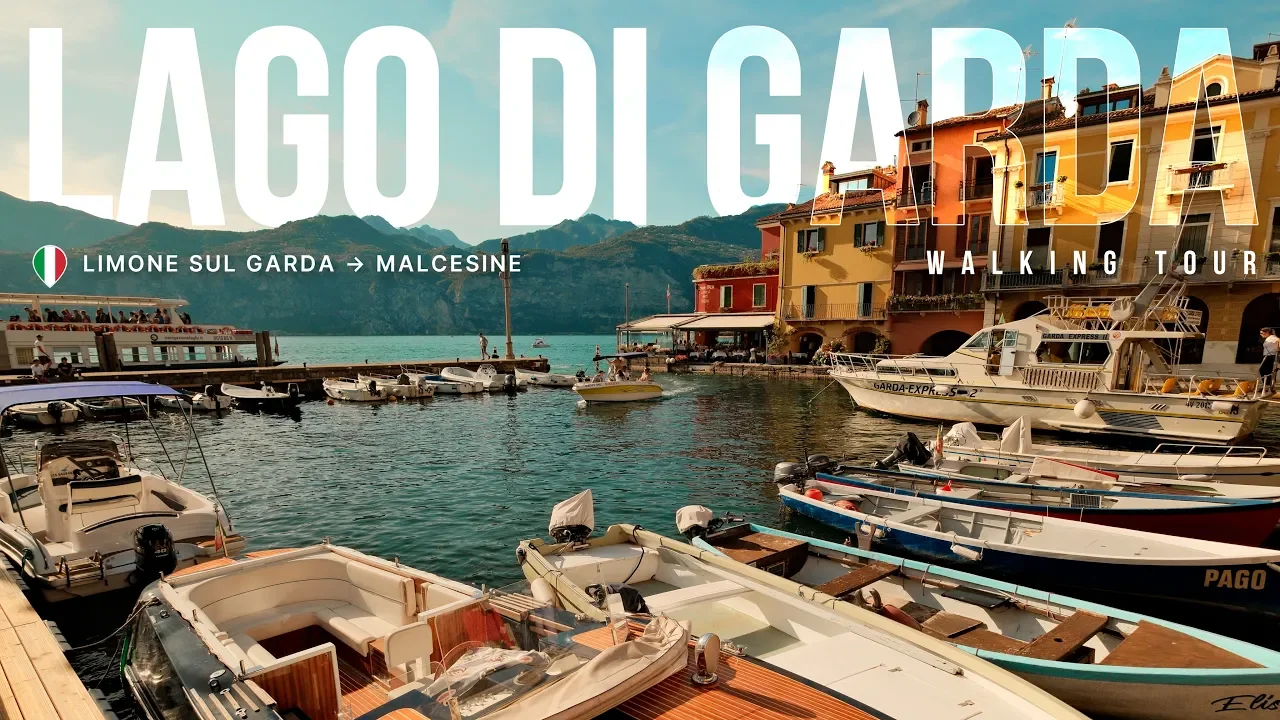 Boats docked at a harbor in Lago di Garda with colorful buildings and mountains in the background, part of a walking tour from Limone sul Garda to Malcesine.