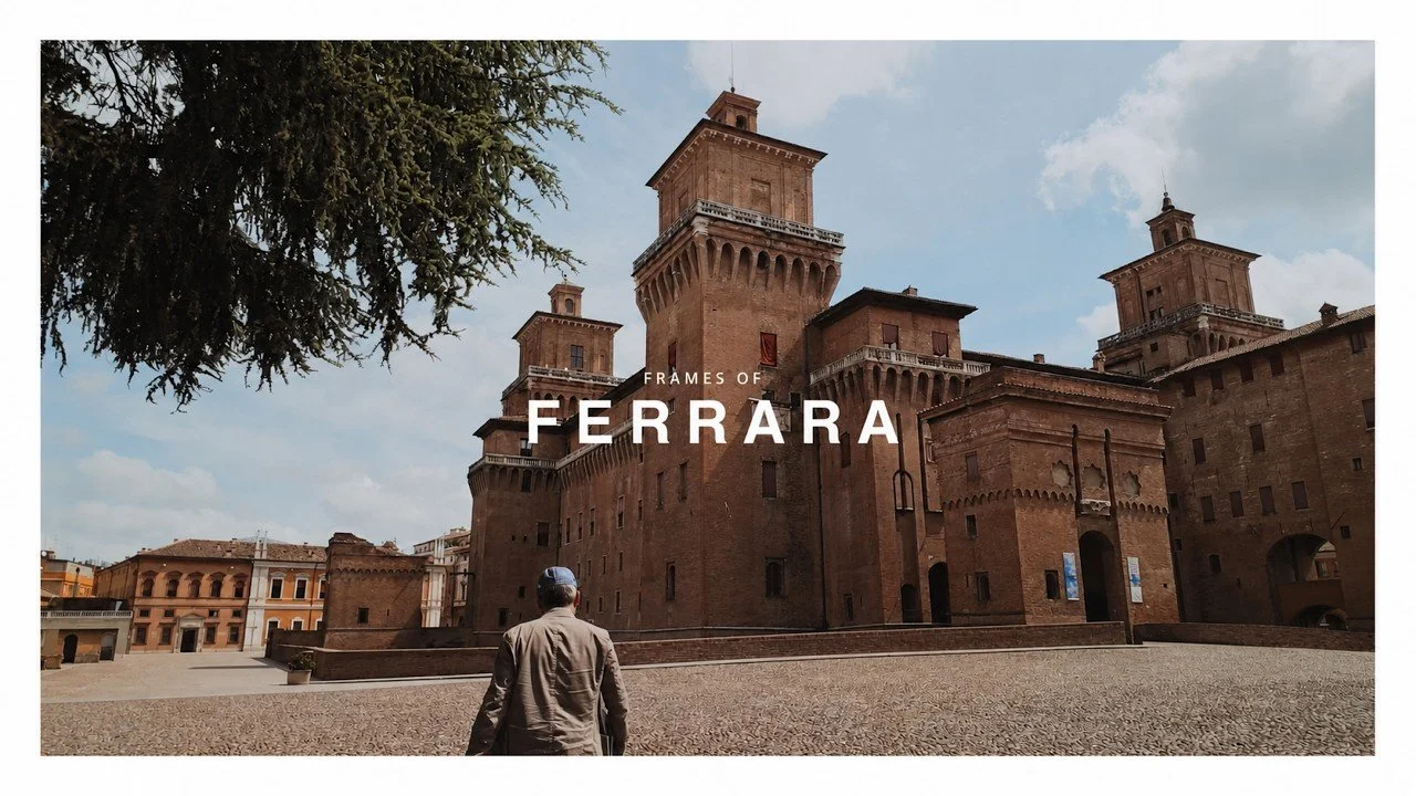 A man walking towards a large medieval castle made of red brick, with towers and battlements, under a partly cloudy sky in Ferrara, Italy.