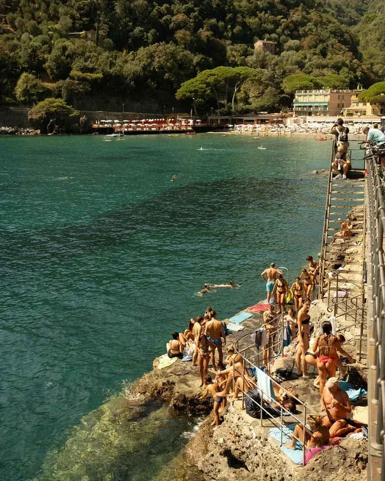 People in swimsuits relaxing on rocks and towels by the water's edge on a sunny day, with some swimming in the water and others standing or sitting near the railing along the shore