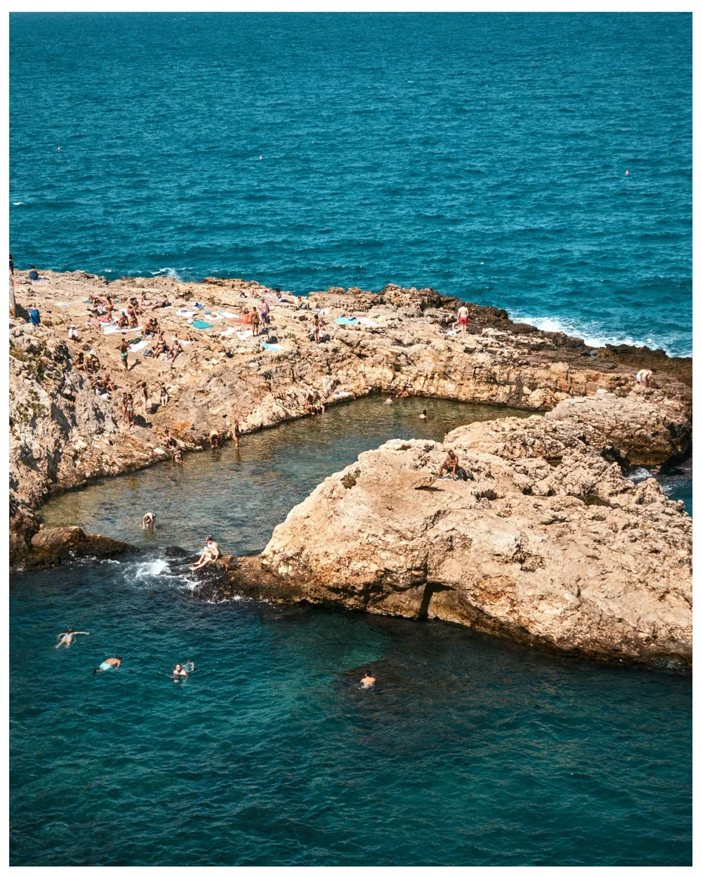 People relaxing and swimming on rocky coastline near a small natural pool, with expansive ocean in the background.