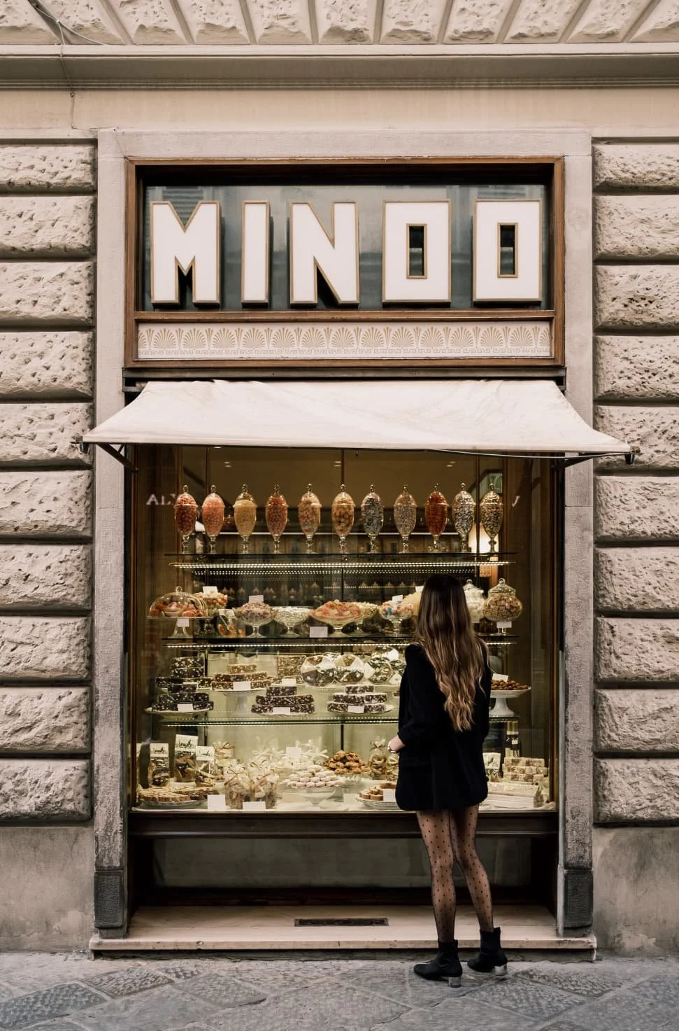 A woman with long hair, wearing a black jacket, polka dot tights, and black boots, stands outside a bakery named 'MINDO,' looking at a display of desserts and chocolates through a glass window.