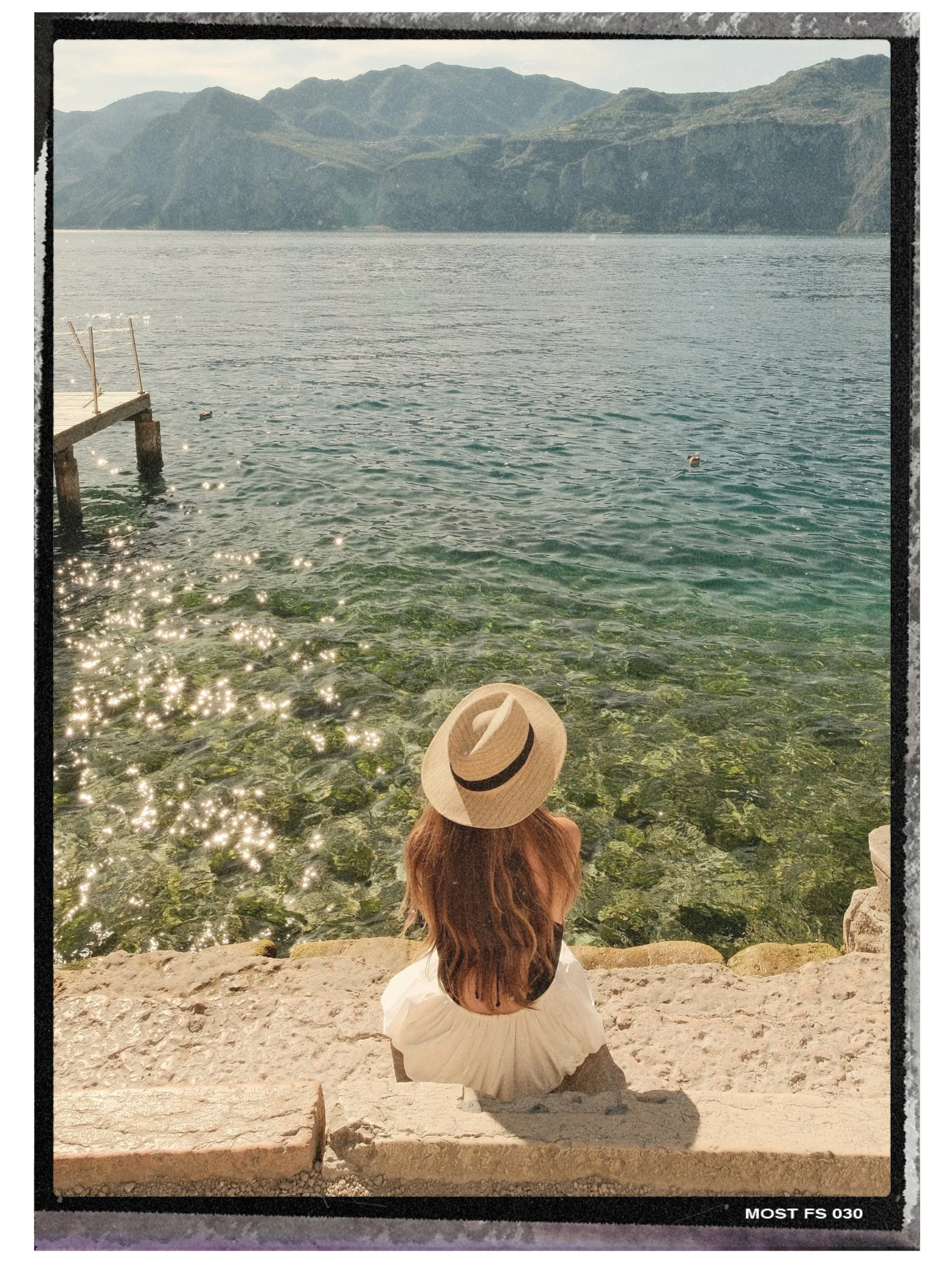 A woman with long hair and a hat sitting on rocks by a body of water, looking at mountains in the distance with sunlight reflecting off the water.