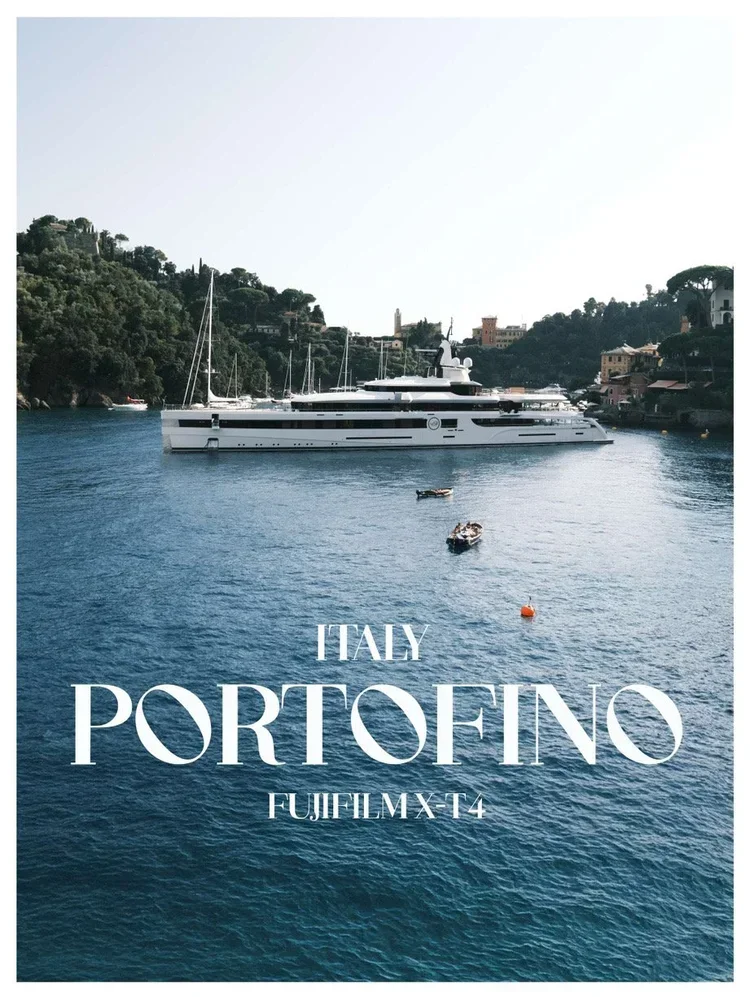 A large white yacht in a harbor with buildings and lush green hills in the background, with smaller boats floating nearby, under a clear sky. The text reads "Italy Portofino Fujifilm X-T4."