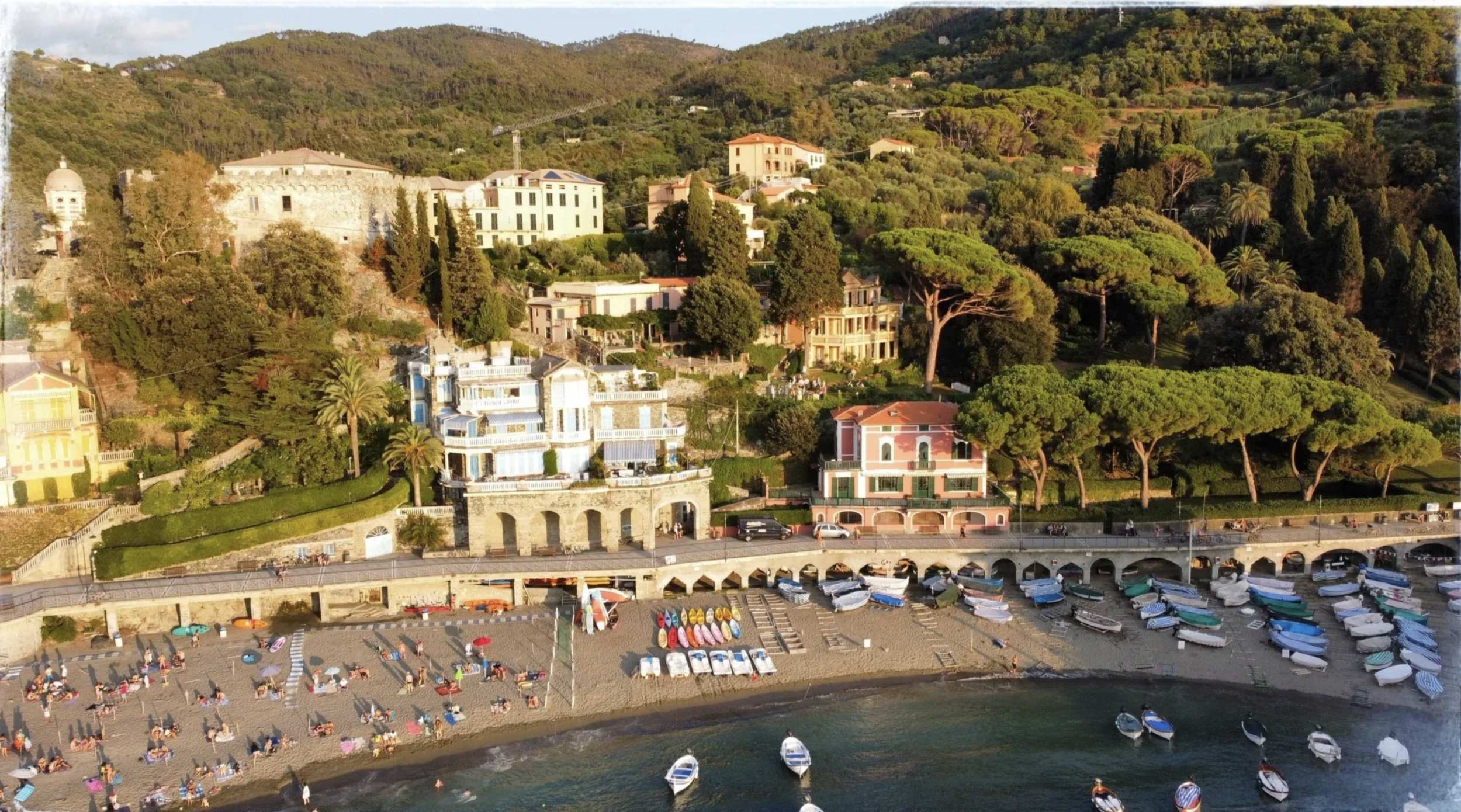 A coastal hillside with colorful houses, lush green trees, and a marina with boats, near a beach filled with sunbathers and umbrellas.
