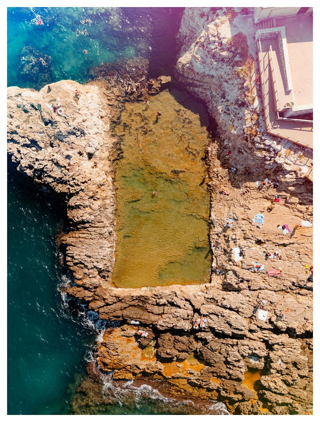 An aerial view of a rocky coastal area with a small, rectangular swimming pool filled with water, surrounded by people lounging and sunbathing on the rocks. The ocean is visible on the left side of the image.