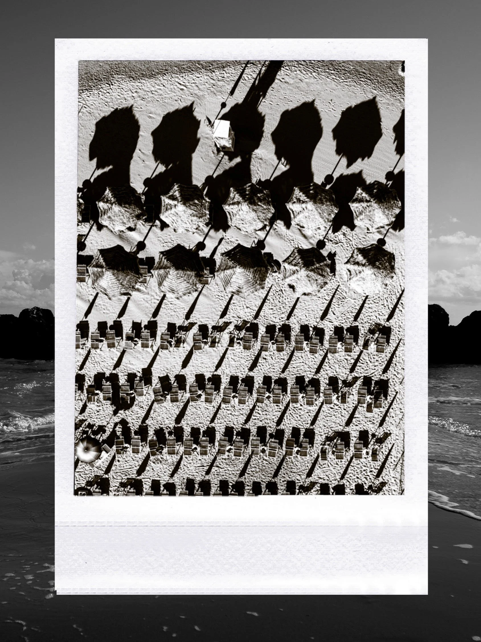 A black and white aerial photograph of a beach with umbrellas and beach chairs arranged in rows. The photo appears to be a print or a photograph of a photograph, with the background showing ocean waves and a cloudy sky.