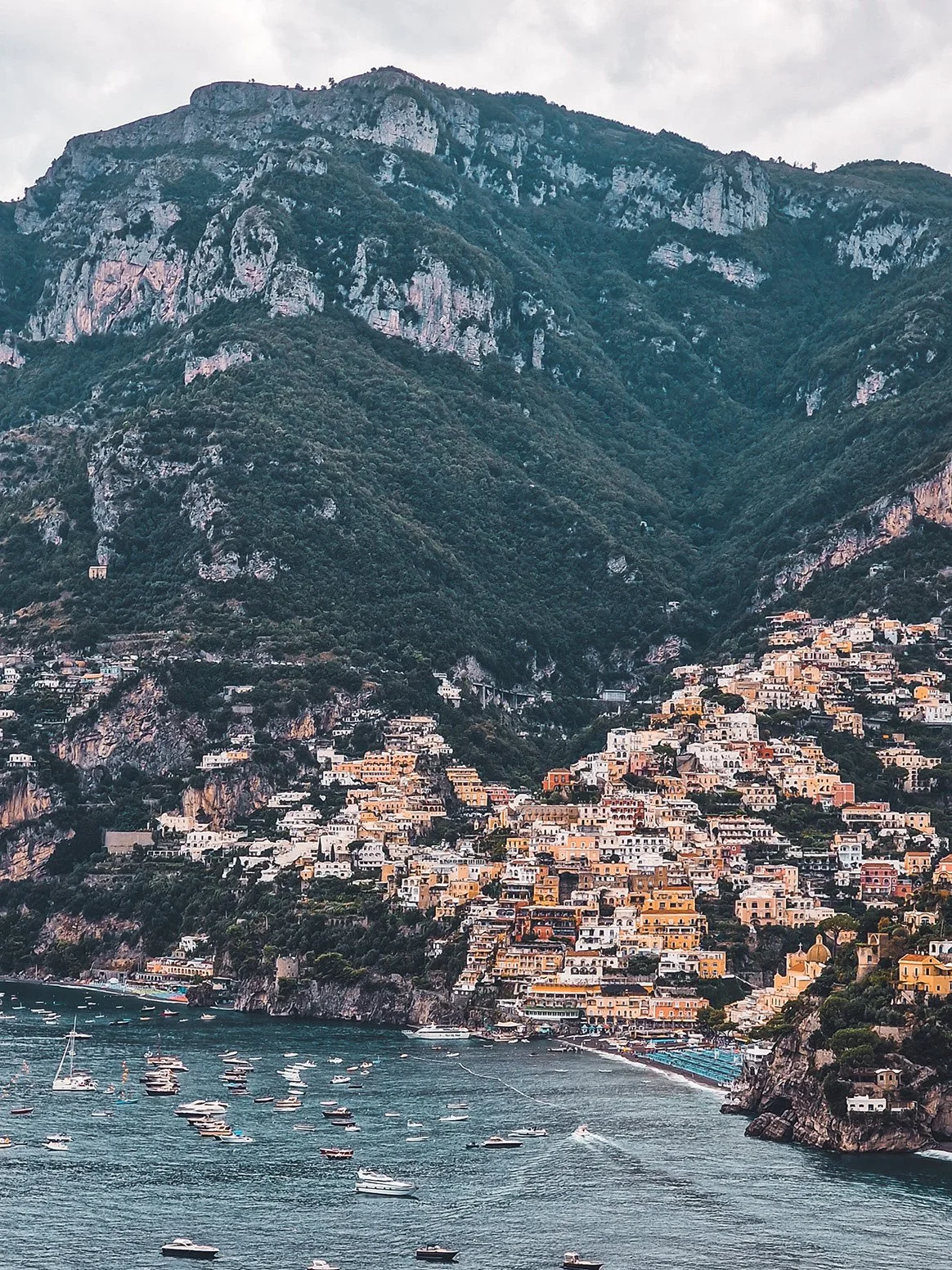 Coastal village with colorful buildings built on a steep hillside next to a large mountain, overlooking a water area with boats.
