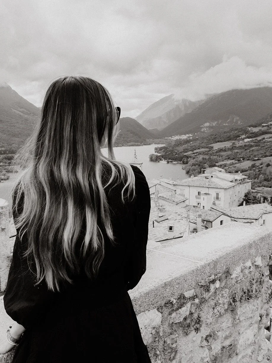 A woman with long wavy hair and sunglasses looking over a stone wall at a small town, river, mountains, and cloudy sky in the distance.