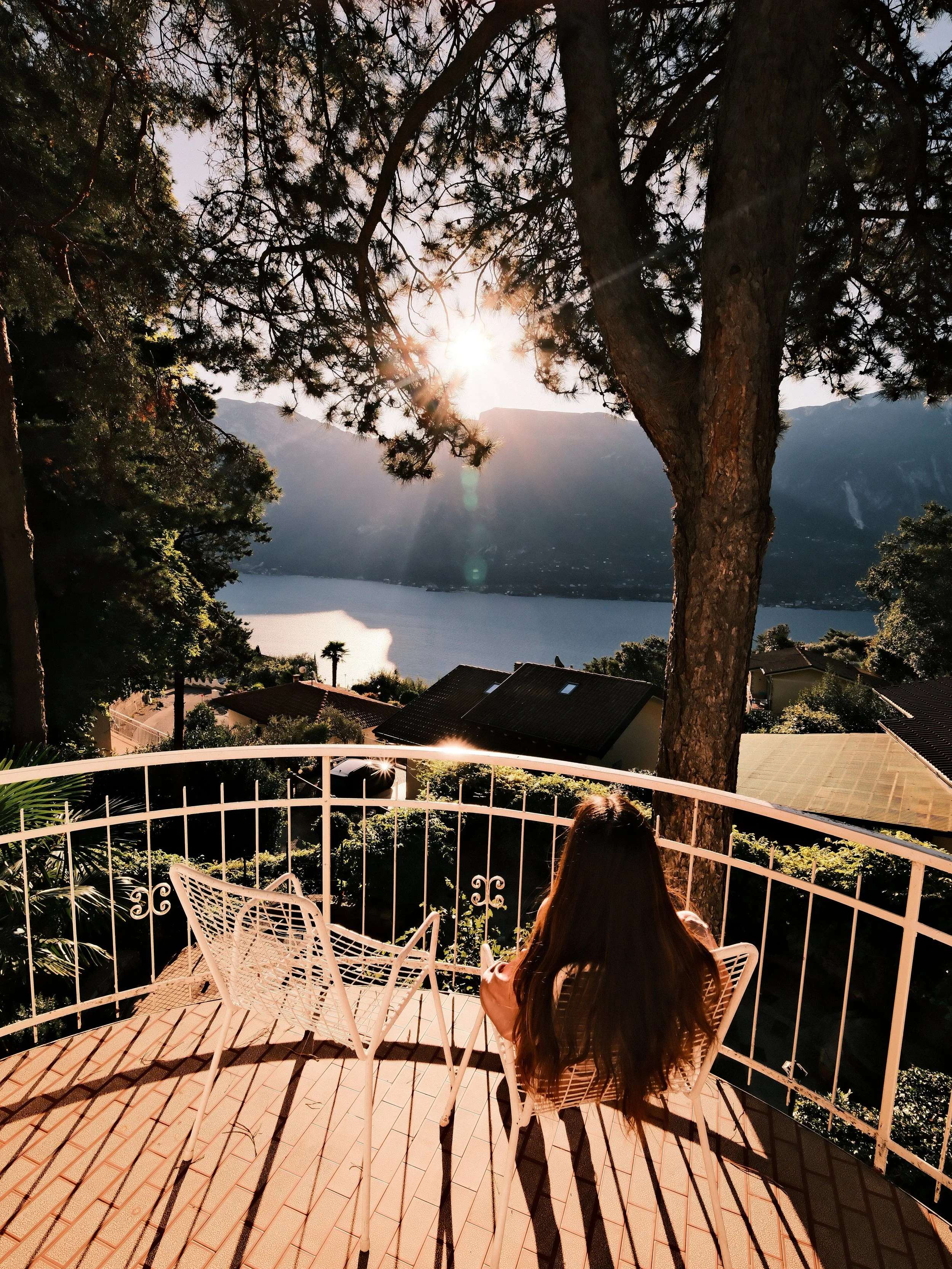 A woman with long brown hair sitting on a white metal chair on a terrace, overlooking a lake and mountain landscape, with the setting sun shining through trees in the background.