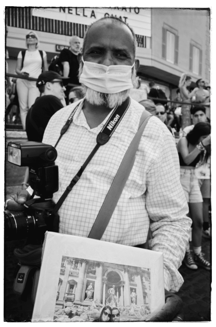 A man wearing a face mask and a Nikon camera strap around his neck holding a photo or printout, standing among a crowd of people in front of a building.