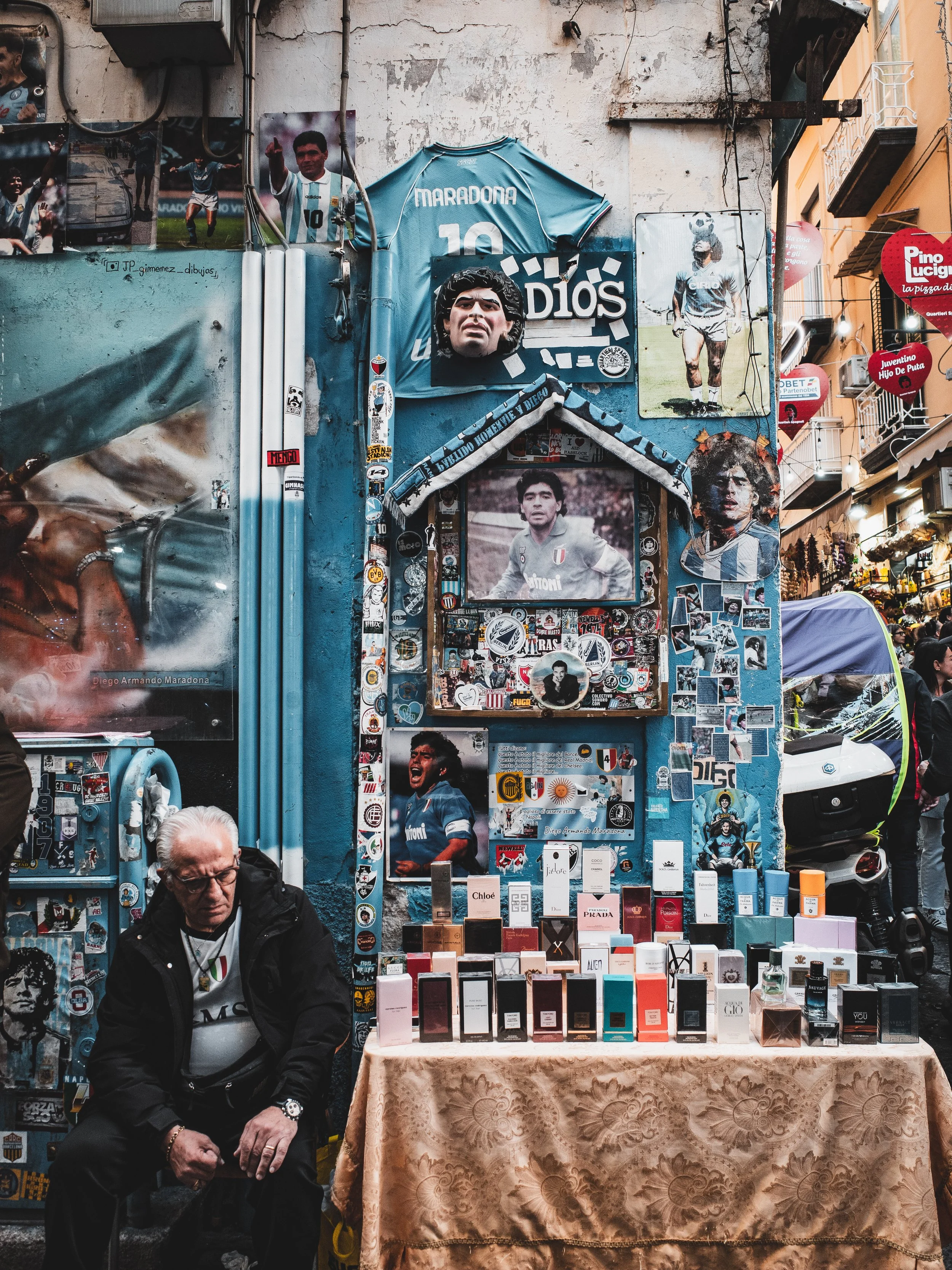 Street market stall decorated with posters, photographs, stickers, and memorabilia of famous soccer player Diego Armando Maradona, including a blue Maradona jersey, a mask, and various phone cases and perfumes displayed on a table. A man with glasses