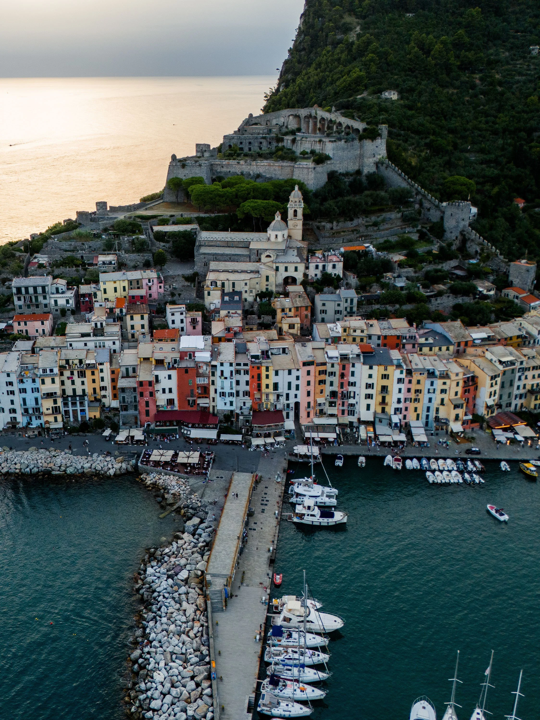 Aerial view of a coastal town with colorful buildings, a pier with yachts, a castle on a hill, and mountains in the background.