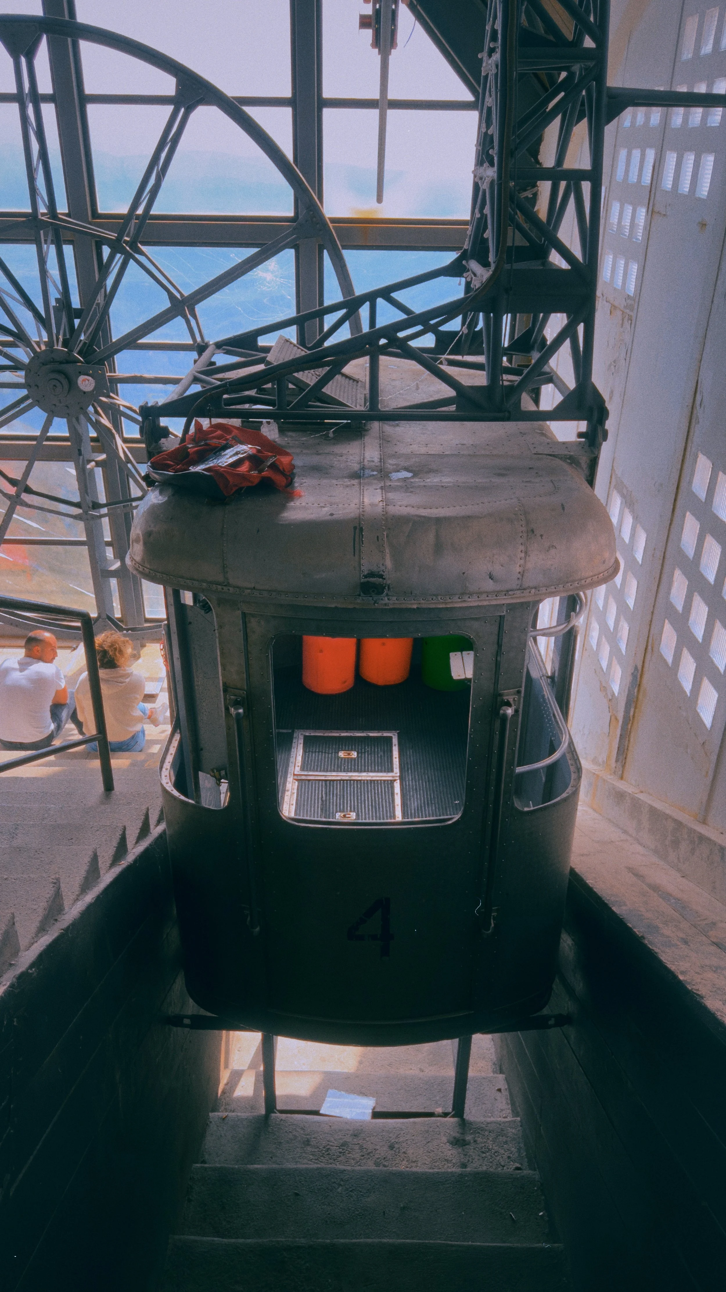 View inside a cable car or gondola lift station, showing the front of the enclosed cabins with three tanks inside, and a structure supporting the cable pulley system above. Two people are sitting on seats outside the station near the cable car.