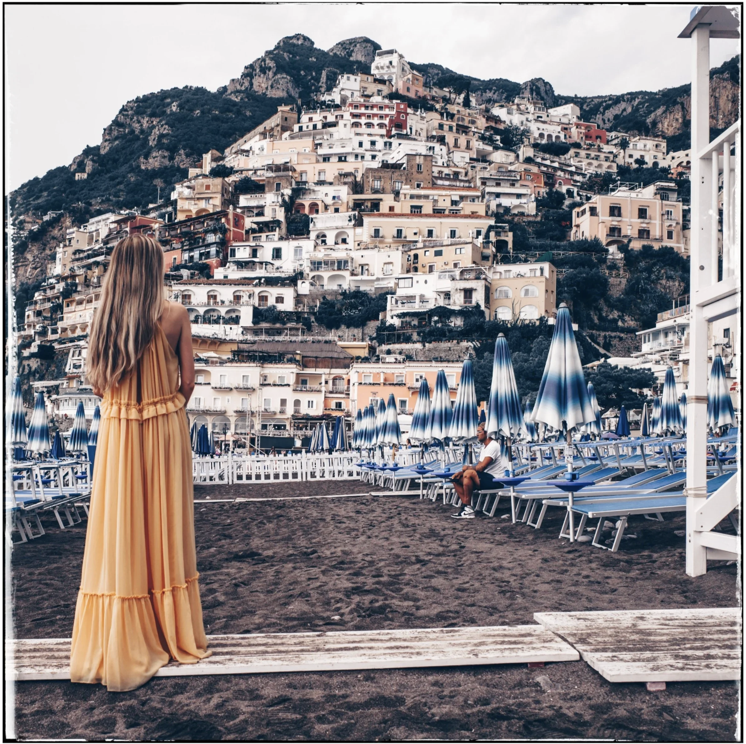 A woman with long blonde hair wearing a yellow dress standing on a beach promenade with a scenic hillside town in the background, featuring colorful houses and buildings stacked on a steep hill.