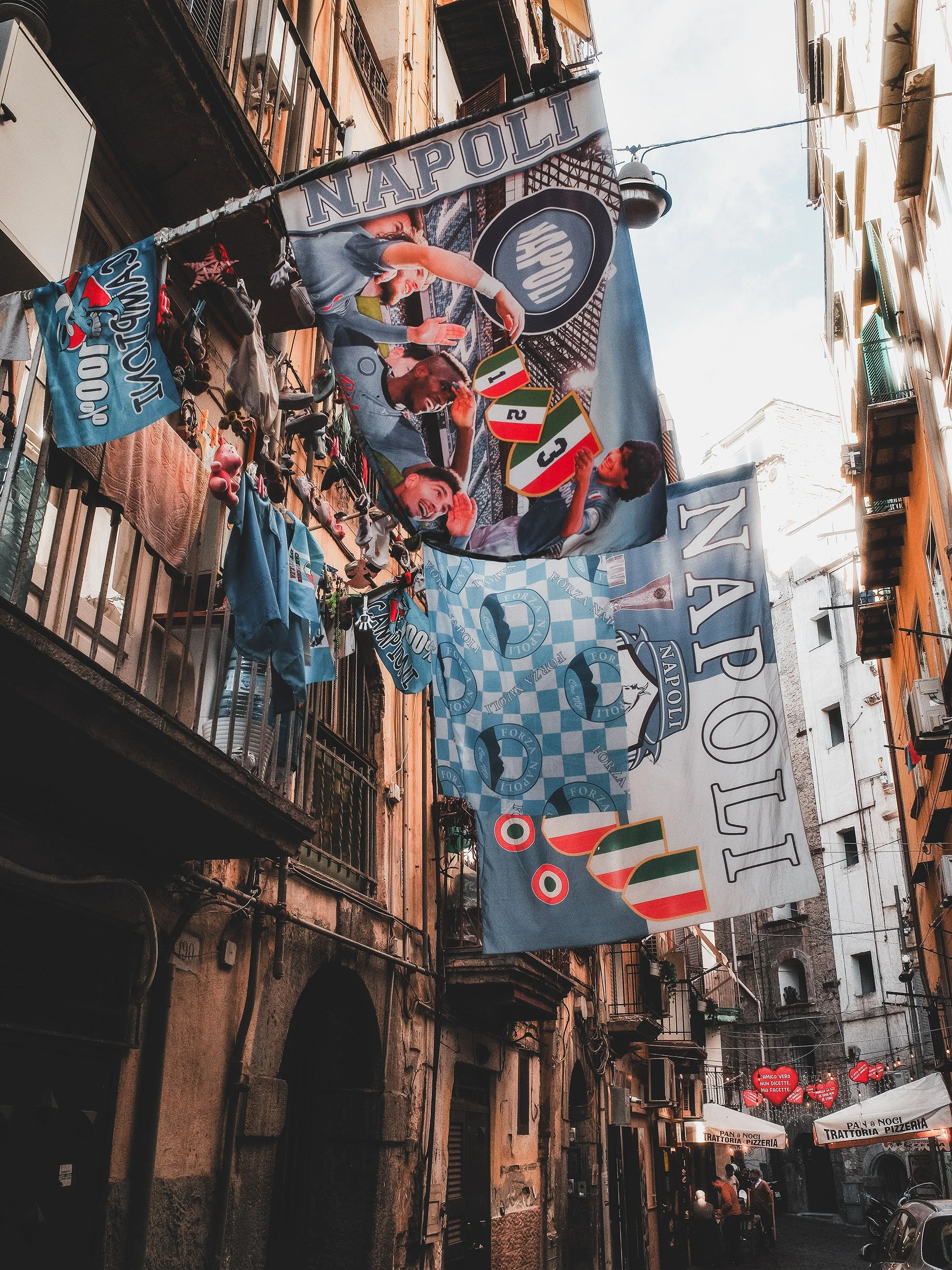City street with banners and hanging objects. Prominent banner displays the word 'Napoli' and features images of football players and team logos. Smaller banner on the left shows a cartoon mouse mascot with the text 'Civitanova.' Street has buildings