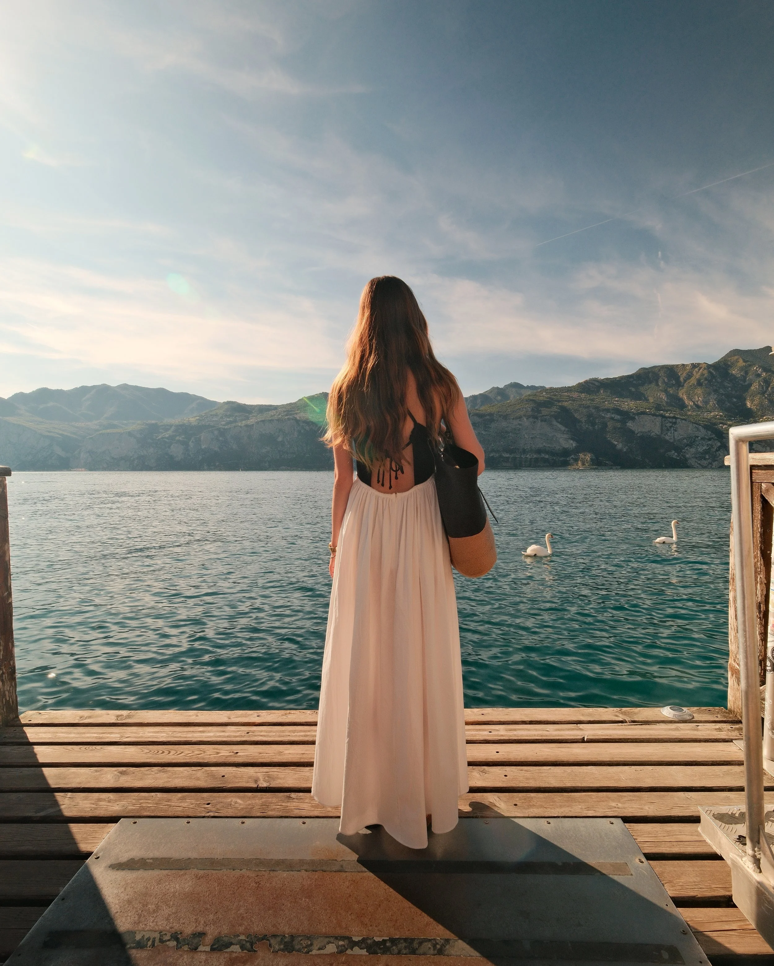 A woman with long hair in a black top and beige skirt standing on a wooden dock, looking at a lake with mountains in the distance, accompanied by two swans in the water, with a clear sky overhead.