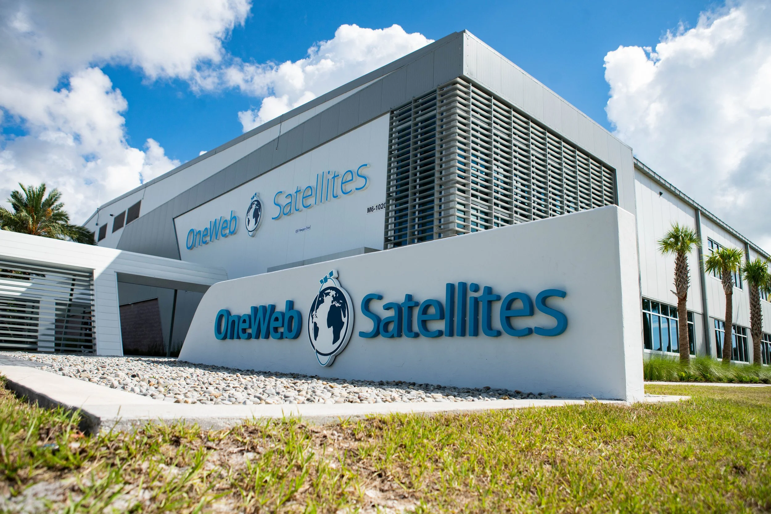 Exterior of a modern building with a sign that reads 'OneWeb Satellites' and a logo of a globe with an orbit ring, under a partly cloudy sky with palm trees nearby.