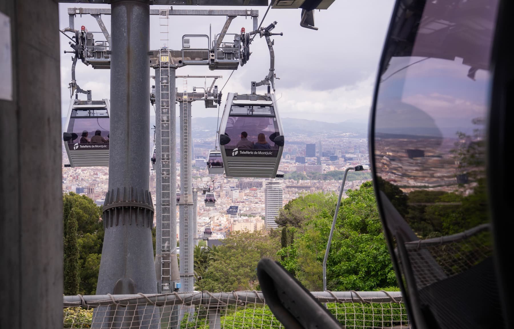 View from inside a Telefèric de Montjuïc gondola looking out at other cabins on the cable line, with the Barcelona cityscape, rooftops, and distant hills below under a cloudy sky.