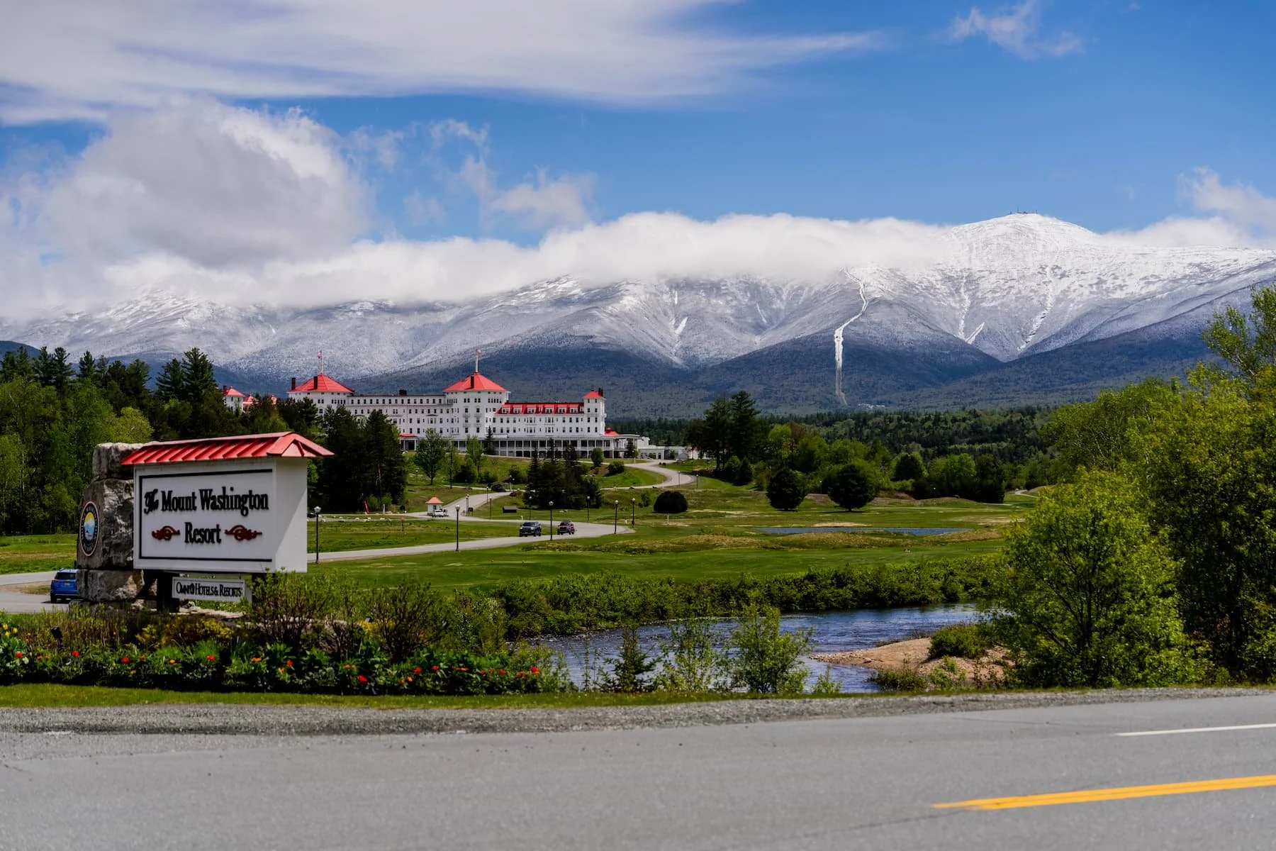 The Mount Washington Resort sign and Omni hotel in Bretton Woods, New Hampshire, with freshly snow-covered White Mountains peaks visible under a blue sky following a late May 2026 snowstorm that dropped 8-10 inches on the summit of Mount Washington.