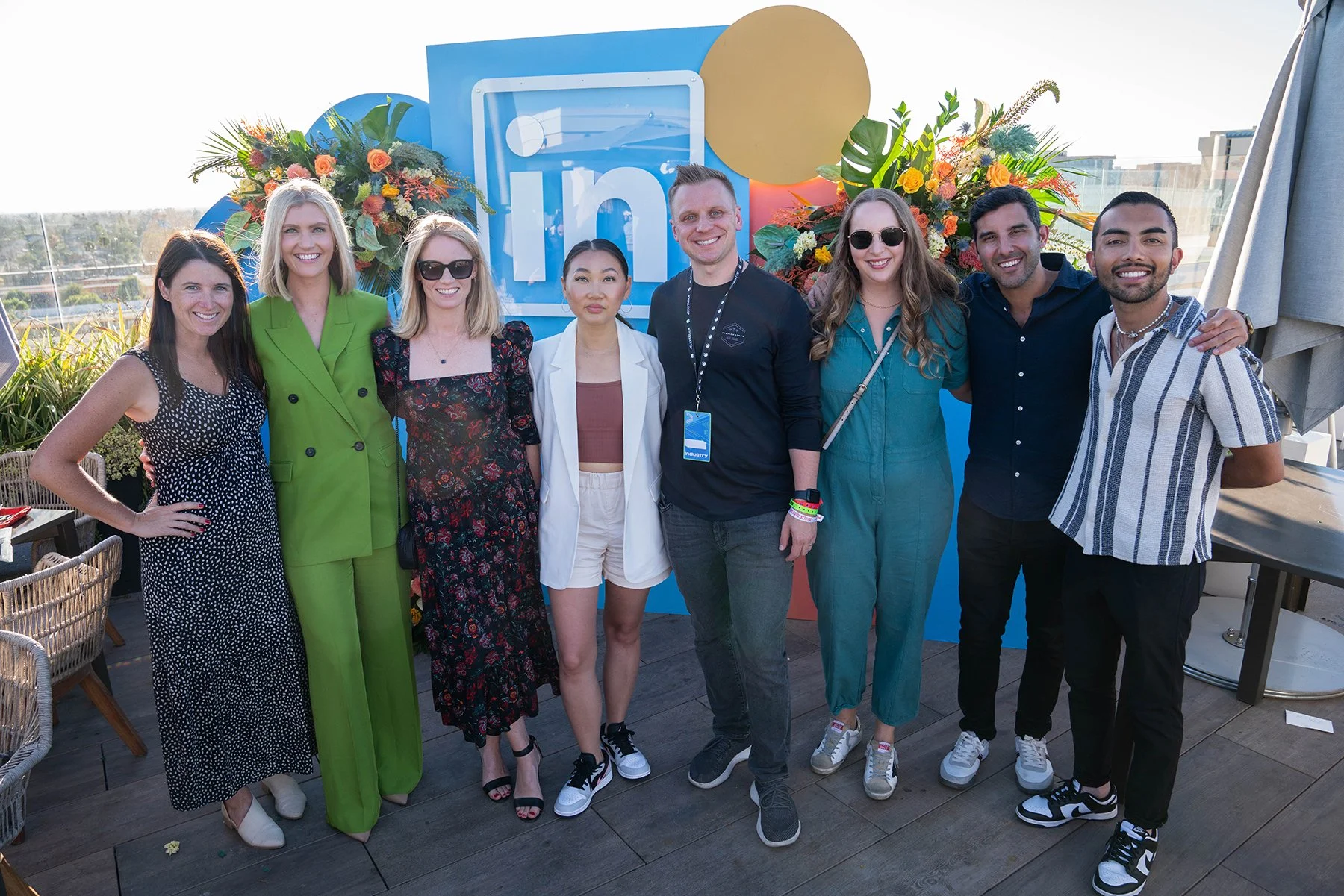 Group of nine diverse people standing together on a rooftop with an 'In' logo and colorful flowers in the background.